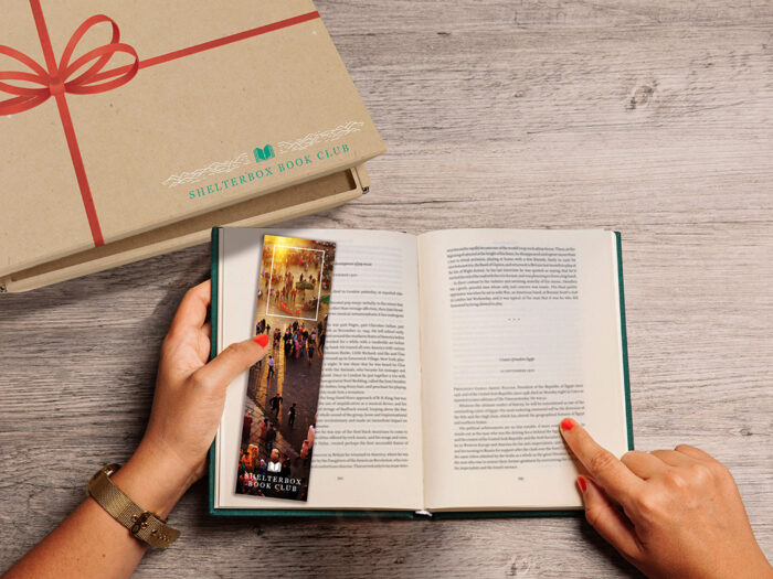 Hands holding open a book with a ShelterBox book club bookmark, and next to a ShelterBox book club branded parcel tied with a red ribbon